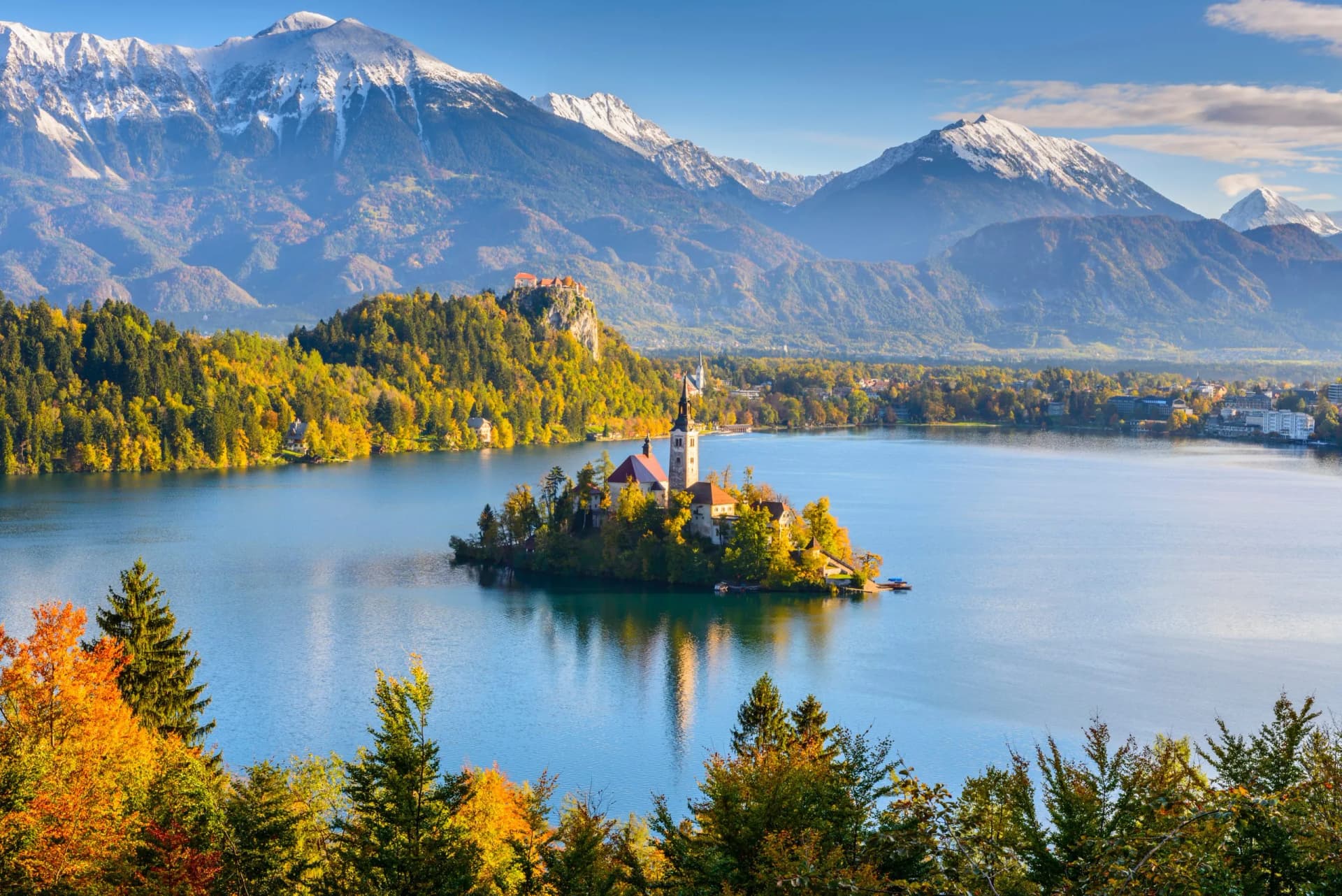 Panoramic view of Lake Bled island church with autumn trees and snow-capped Julian Alps.