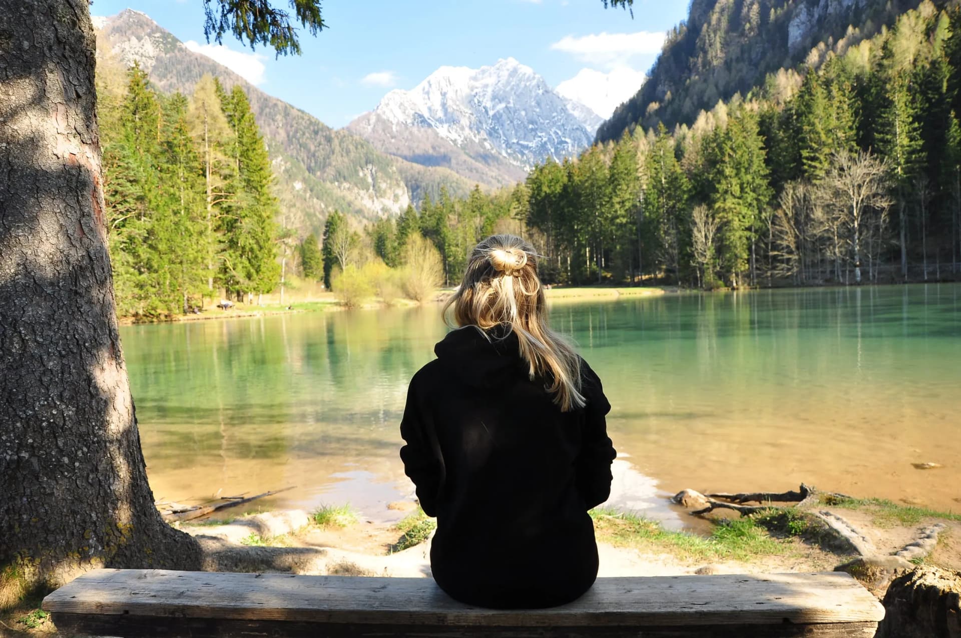 Girl sitting on a bench by Plansarsko Jezero, Slovenia, facing snow-capped mountains.