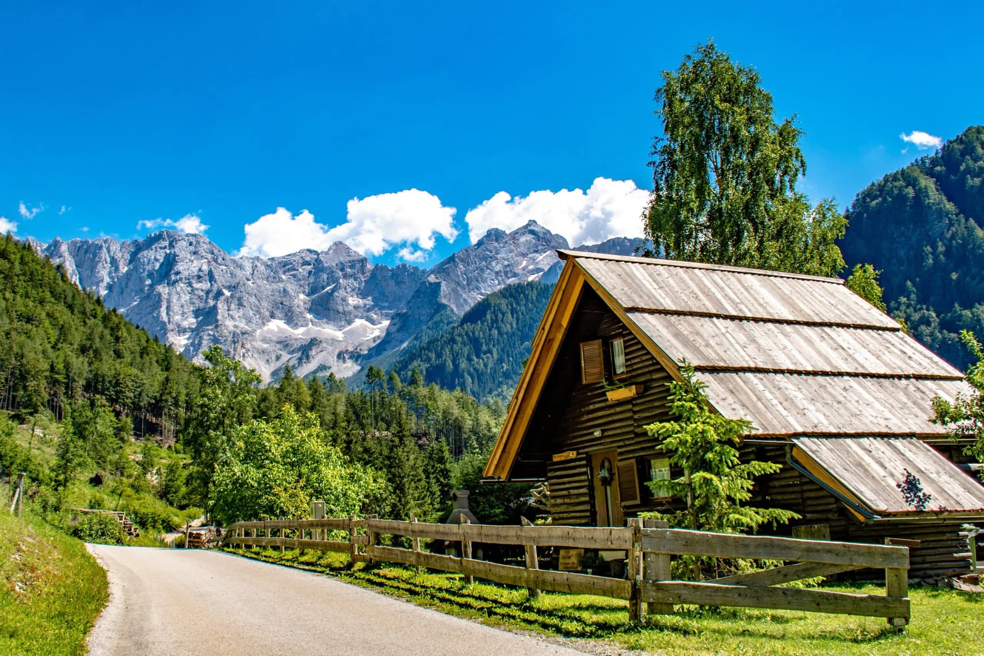 Quaint wooden alpine house near road with snow-capped mountains under blue sky in Zgornje Jezersko.