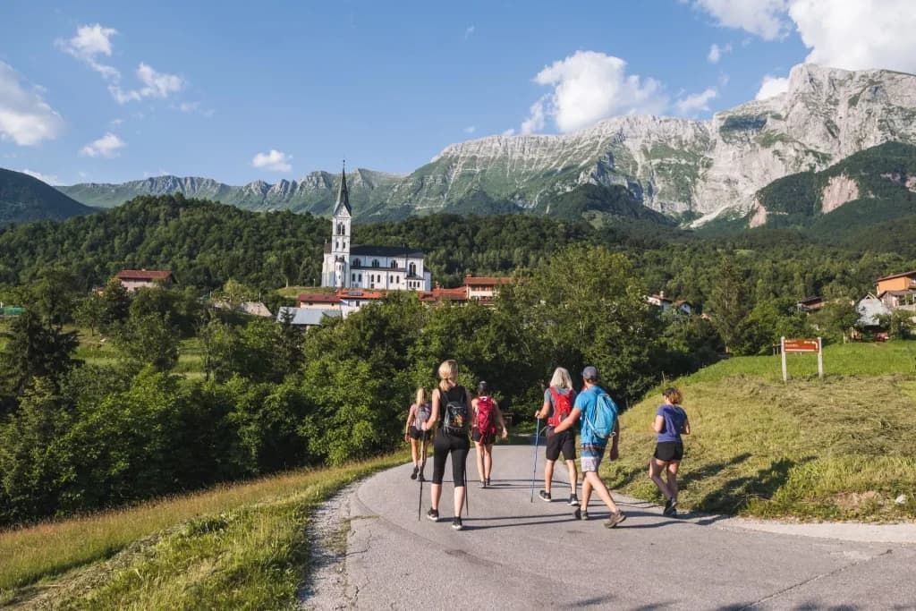 Hikers walking toward a village with a white church under Mt. Krn in Drežnica.