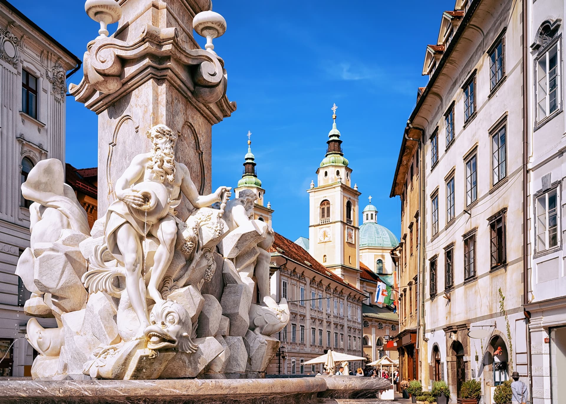 Baroque fountain sculpture with white buildings and church towers in Ljubljana square.
