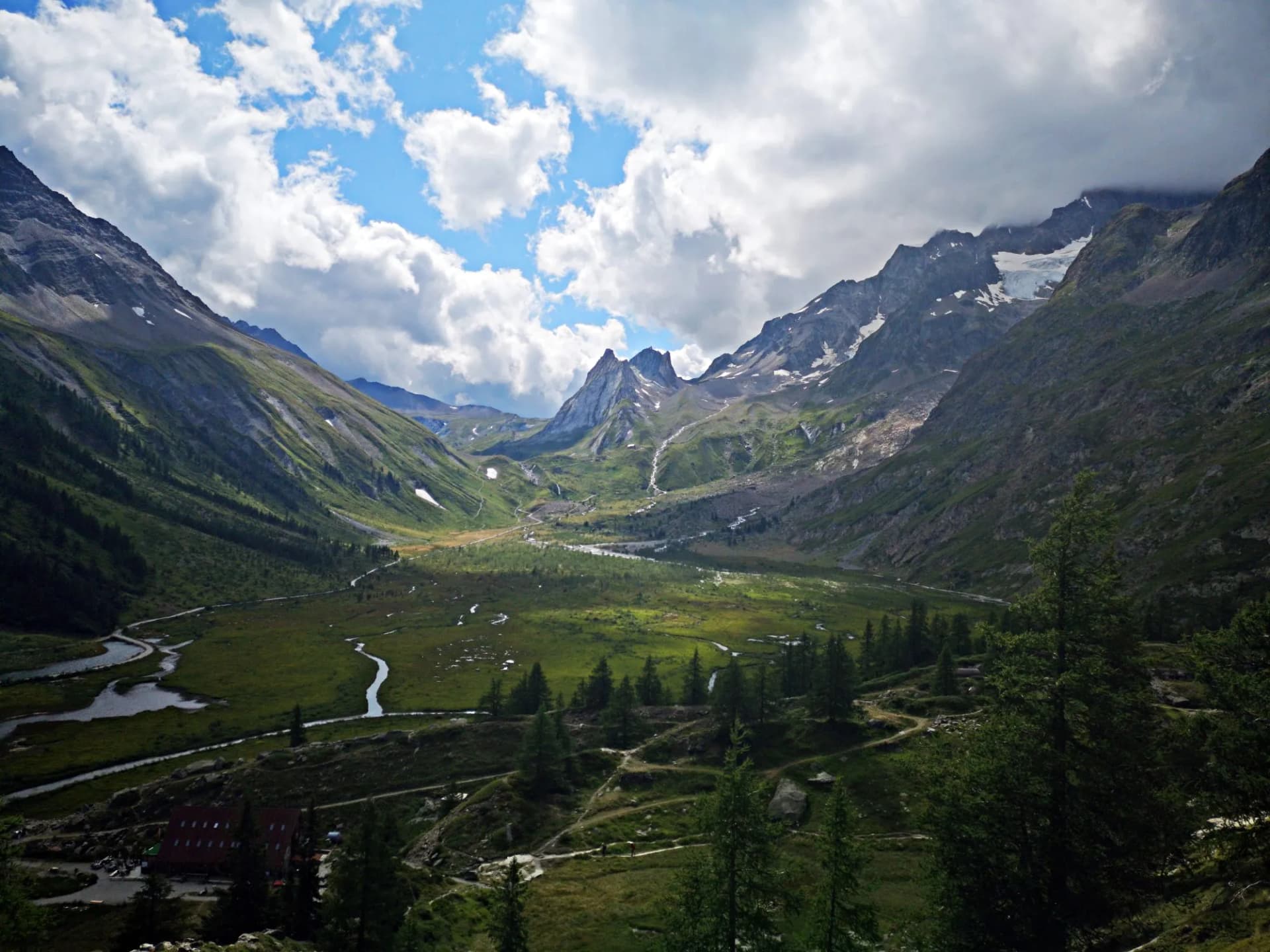 Mountain valley with green slopes, winding stream, and peaks with residual snow under cloudy sky.