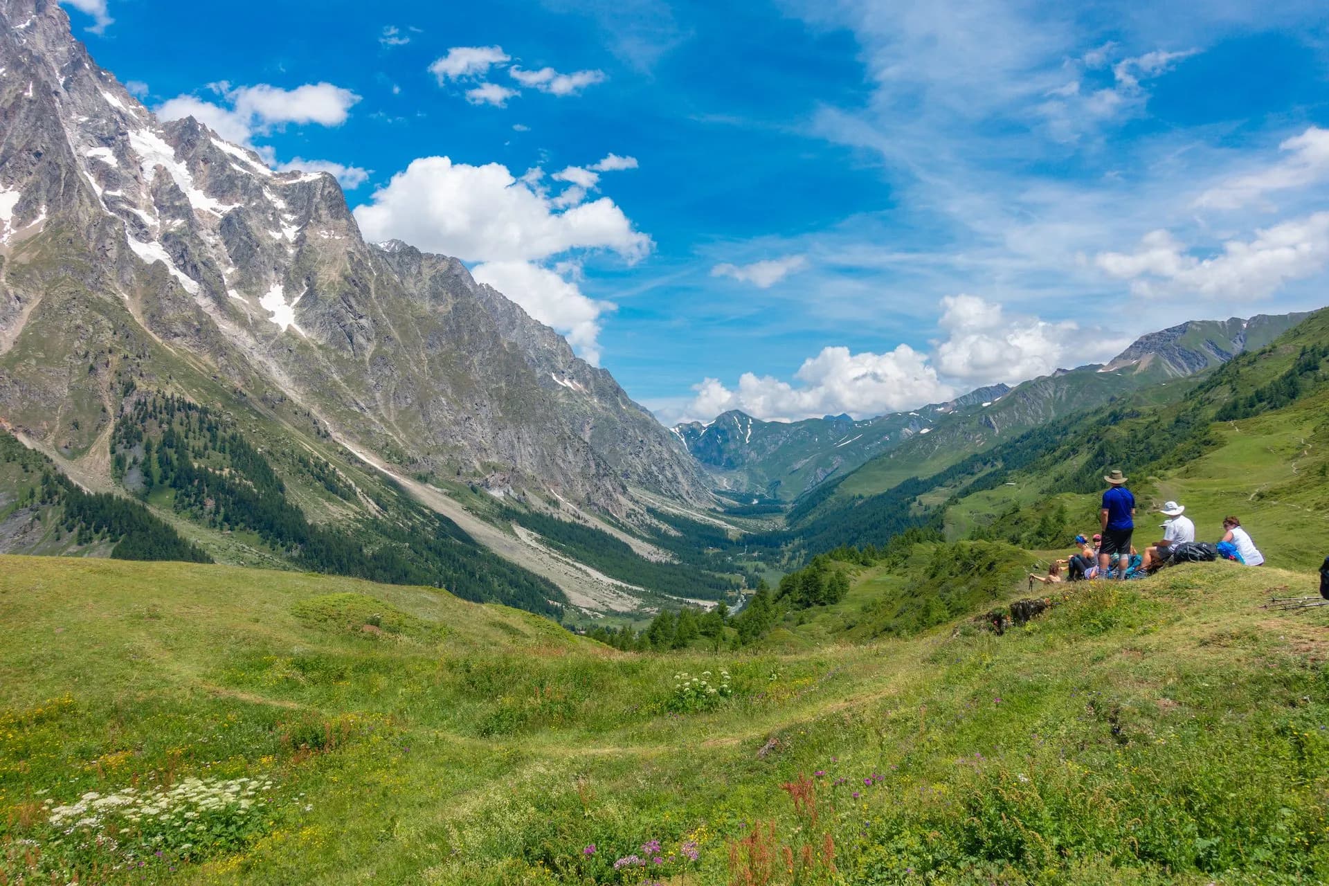 Hikers resting overlooking the vast Val Ferret alpine valley with snow-capped mountains under blue sky.