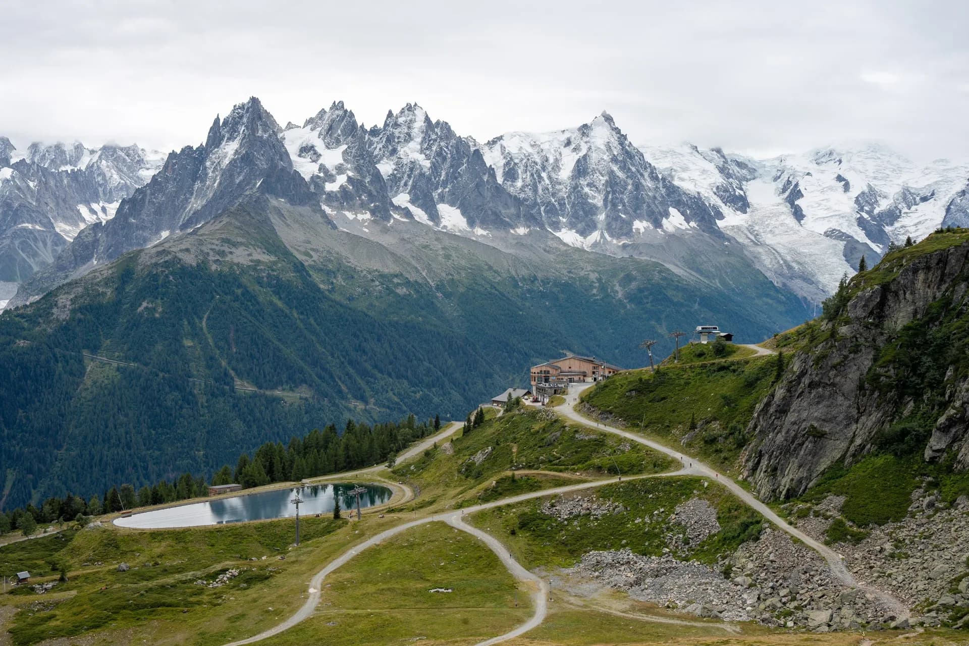 Mountain lodge near reservoir with winding paths below snow-capped peaks, La Flegere.
