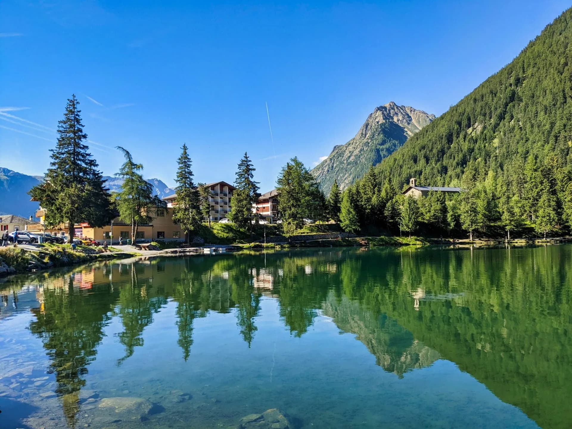 Alpine lake with clear water reflecting mountains, trees, and buildings under a blue sky at Lac de Champex.