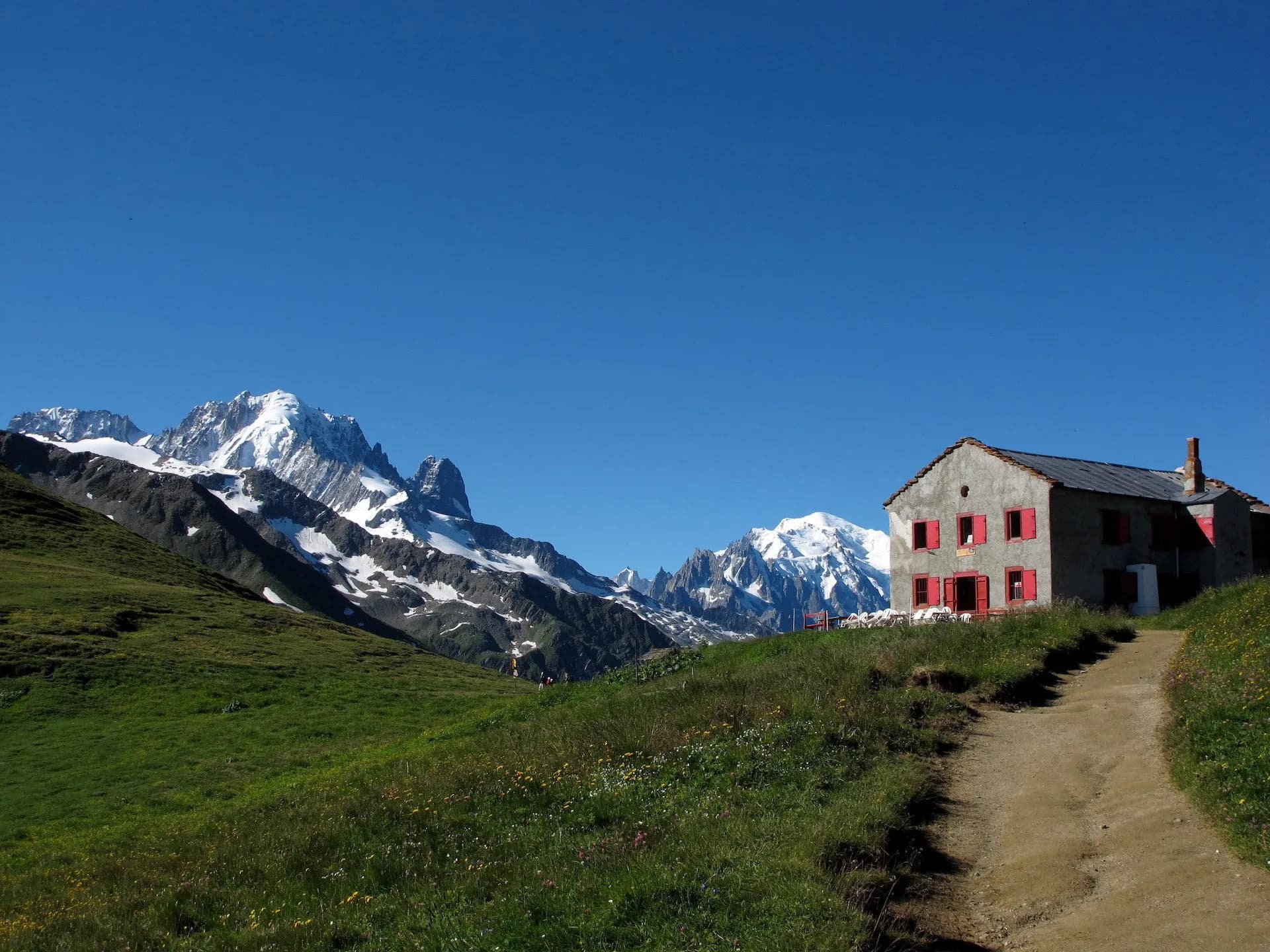 Stone refuge with red shutters on a grassy slope below snow-capped mountains at Col de Balme.