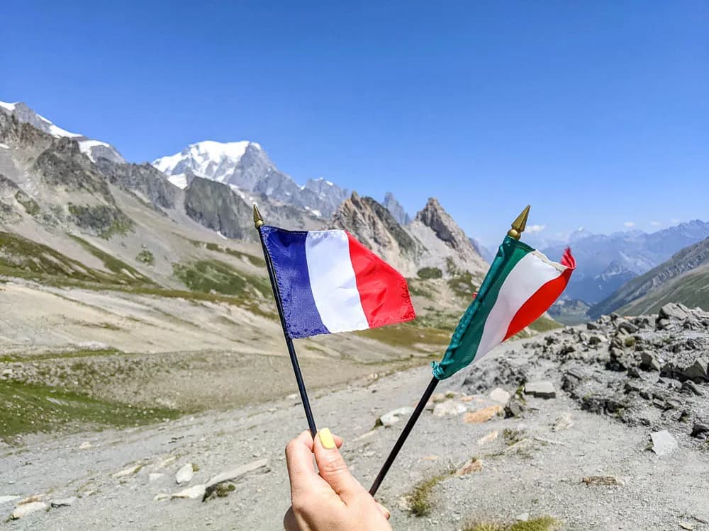 Hand holding French and Italian flags with snow-capped mountains in the background