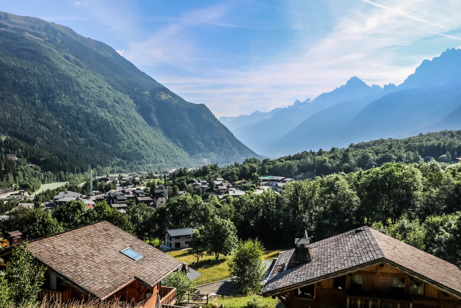 View of Les Houches village nestled in a valley surrounded by steep, forested mountains under a blue sky.
