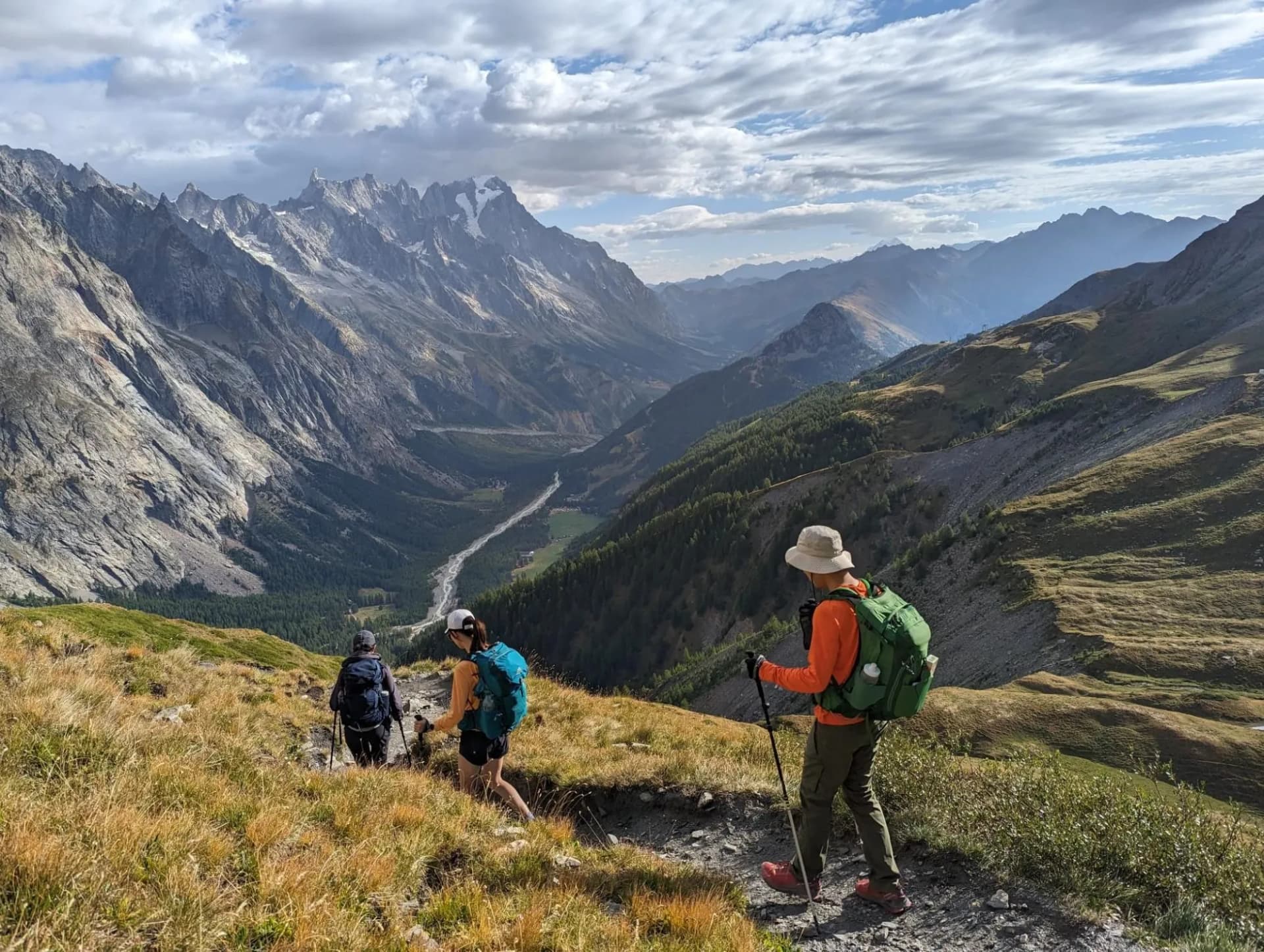 Hikers with backpacks and poles traversing a grassy mountain trail overlooking a deep valley and jagged peaks.