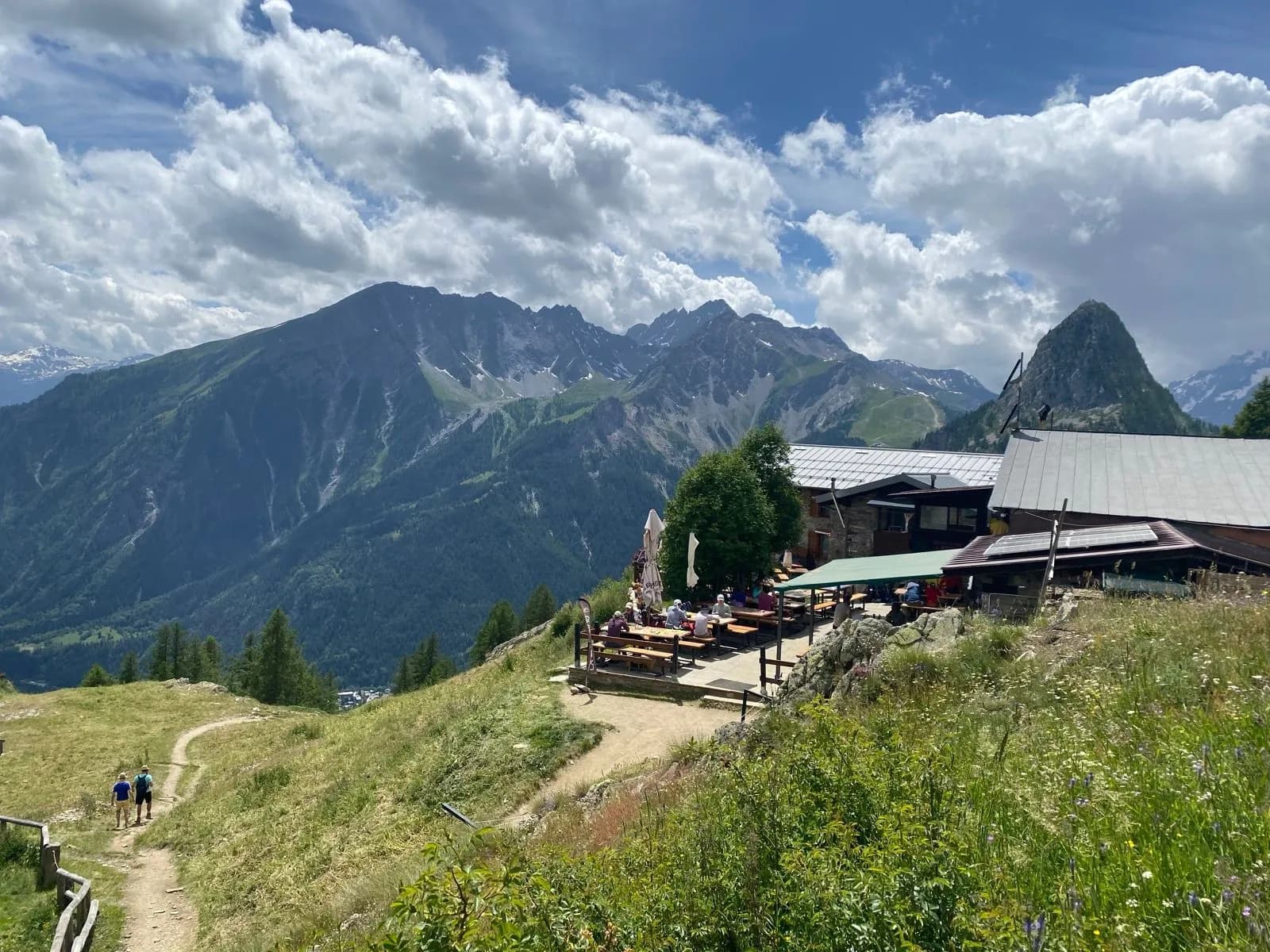 Hikers on trail near mountain refuge with outdoor dining and dramatic peaks under cloudy sky