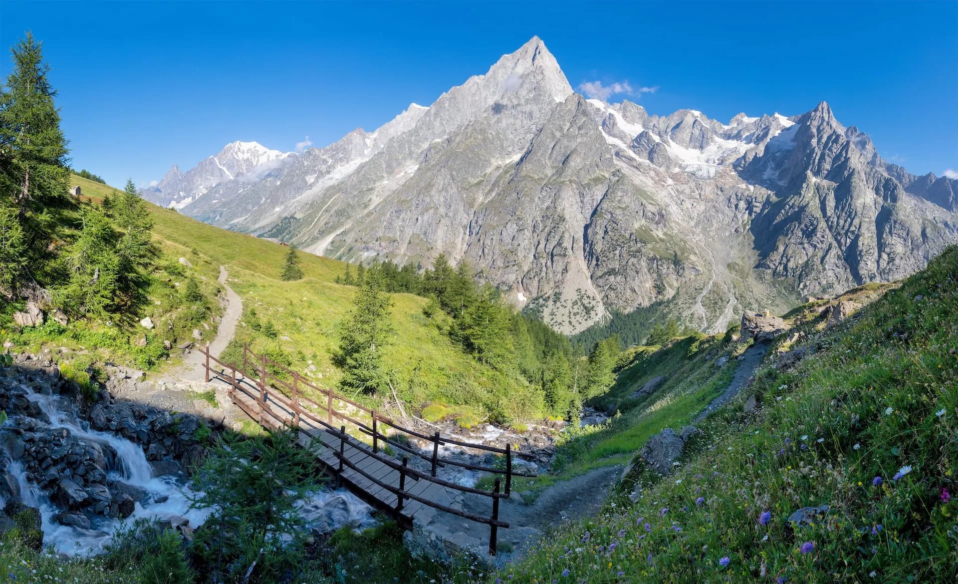 Hiking trail with wooden bridge over stream in Val Ferret valley below snow-capped Jorasses massif.