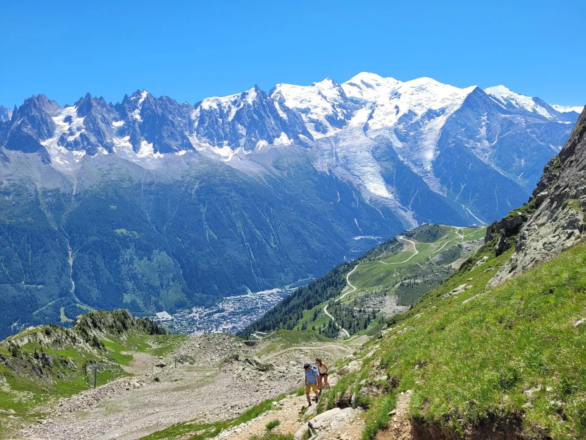 Hikers on trail with view of snow-capped mountains and town below, likely Brevent.