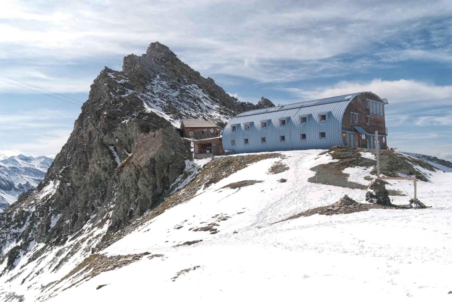 Mountain hut with blue curved roof next to rocky peak on snowy slope with distant peaks.