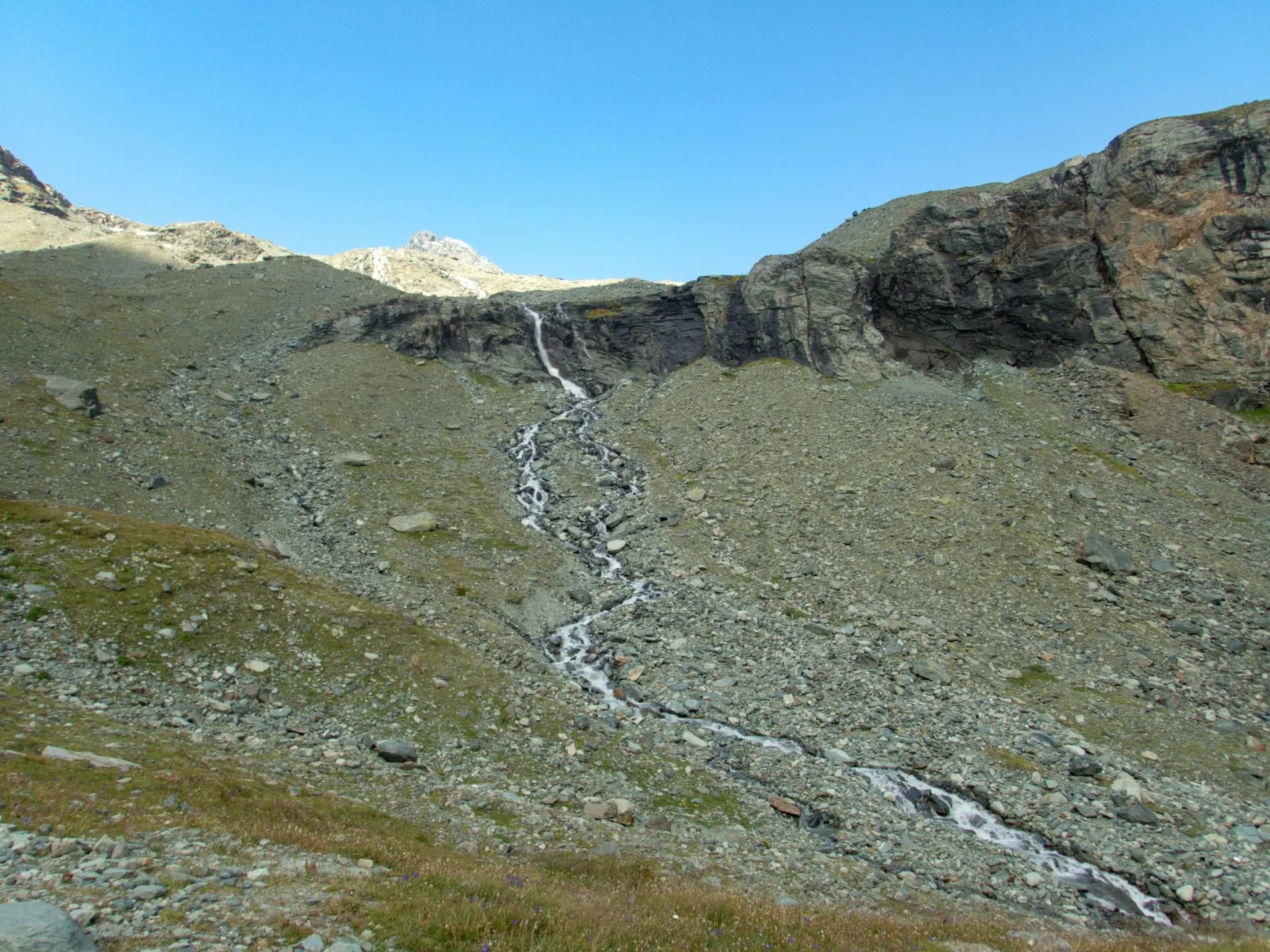Waterfall cascading down a steep, rocky alpine slope under a clear blue sky.