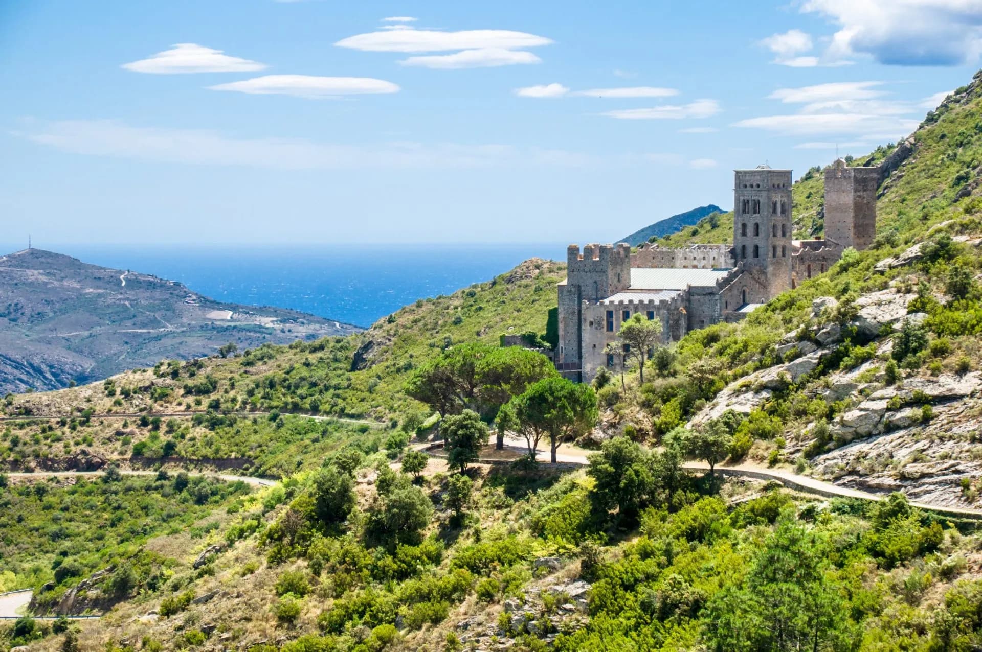 Sant Pere de Rodes monastery on a green, scrub-covered hillside overlooking the blue Mediterranean Sea.