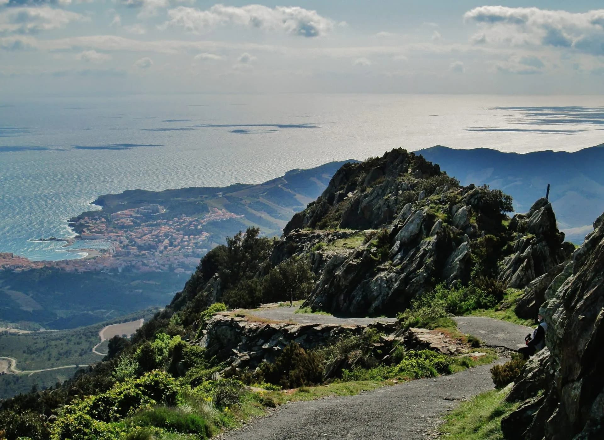 Mountain road overlooking coastal town and sparkling Mediterranean Sea under cloudy sky