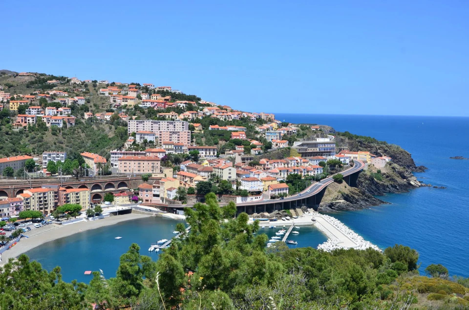 Coastal town with colorful buildings climbing a hill above a harbor and blue sea near a bridge.
