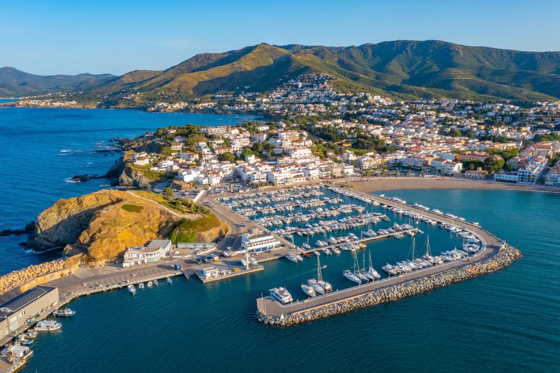 Marina filled with sailboats and yachts on the Mediterranean coast below white hillside town and mountains.
