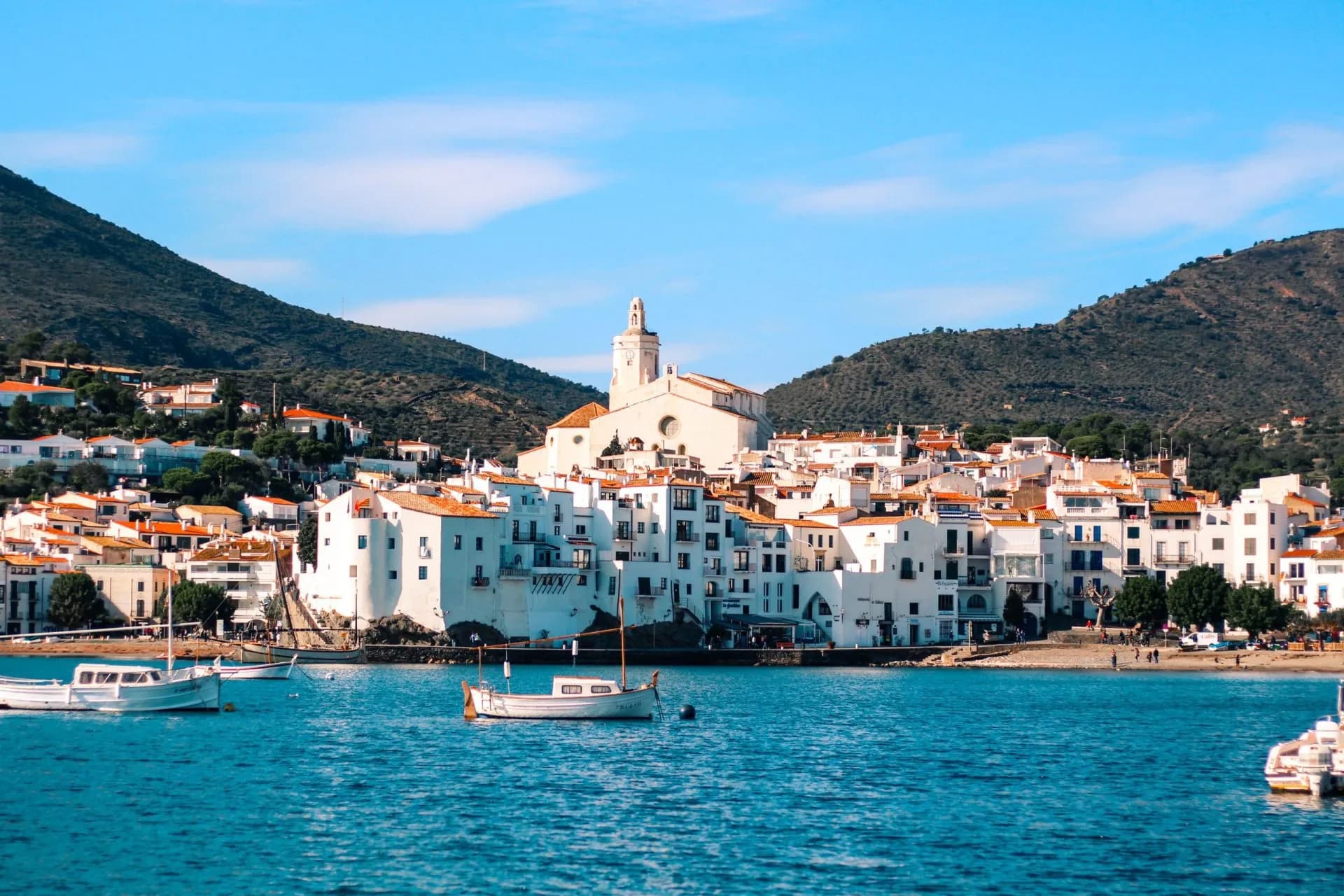 Boats in blue water before white coastal village and green mountains, Cadaqués.