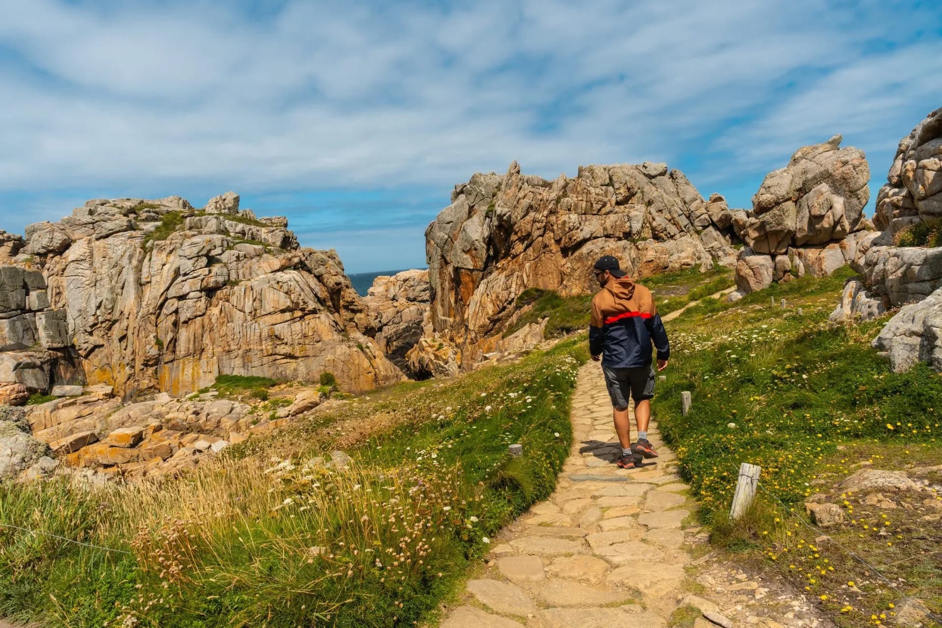Hiking near Le Gouffre de Plougrescant past large coastal rock formations under a blue sky.