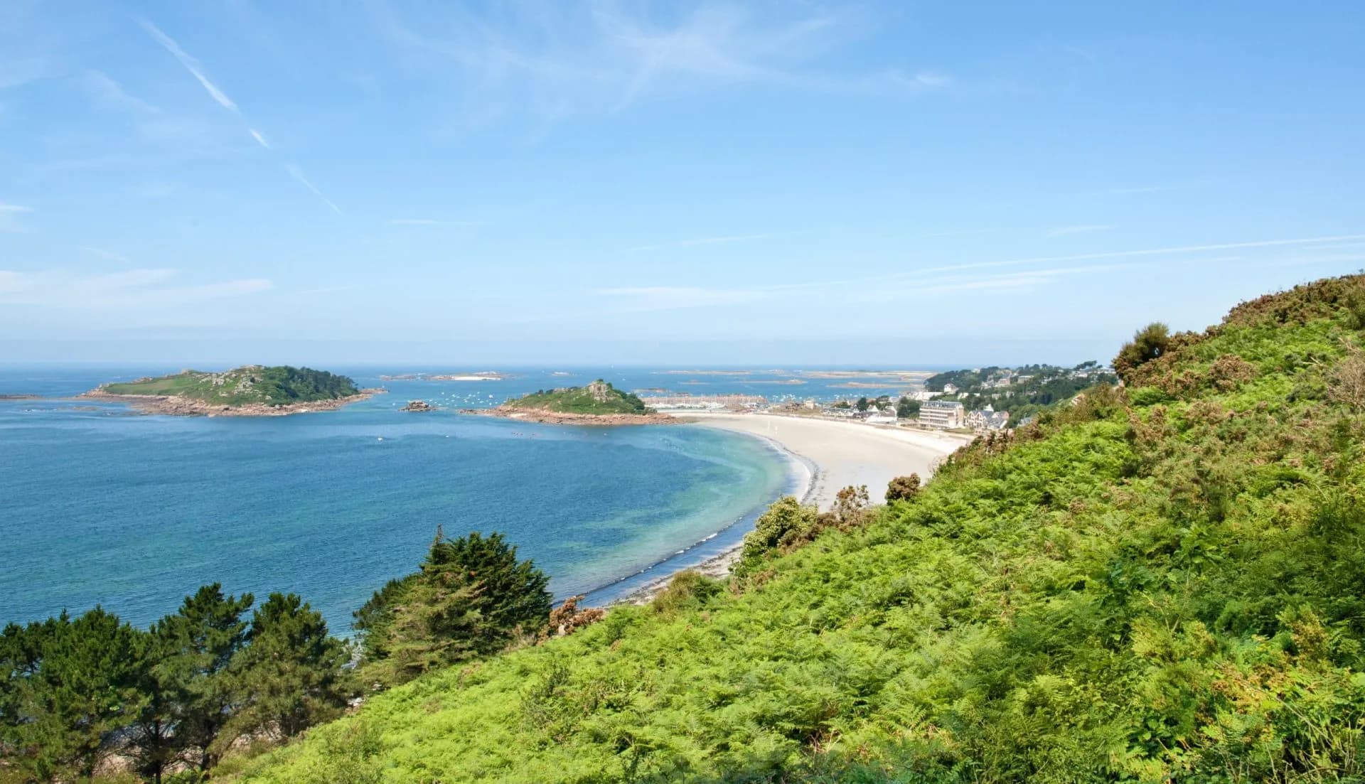 Seaside view of Trebeurden bay with islands, beach, and green hillside under blue sky.
