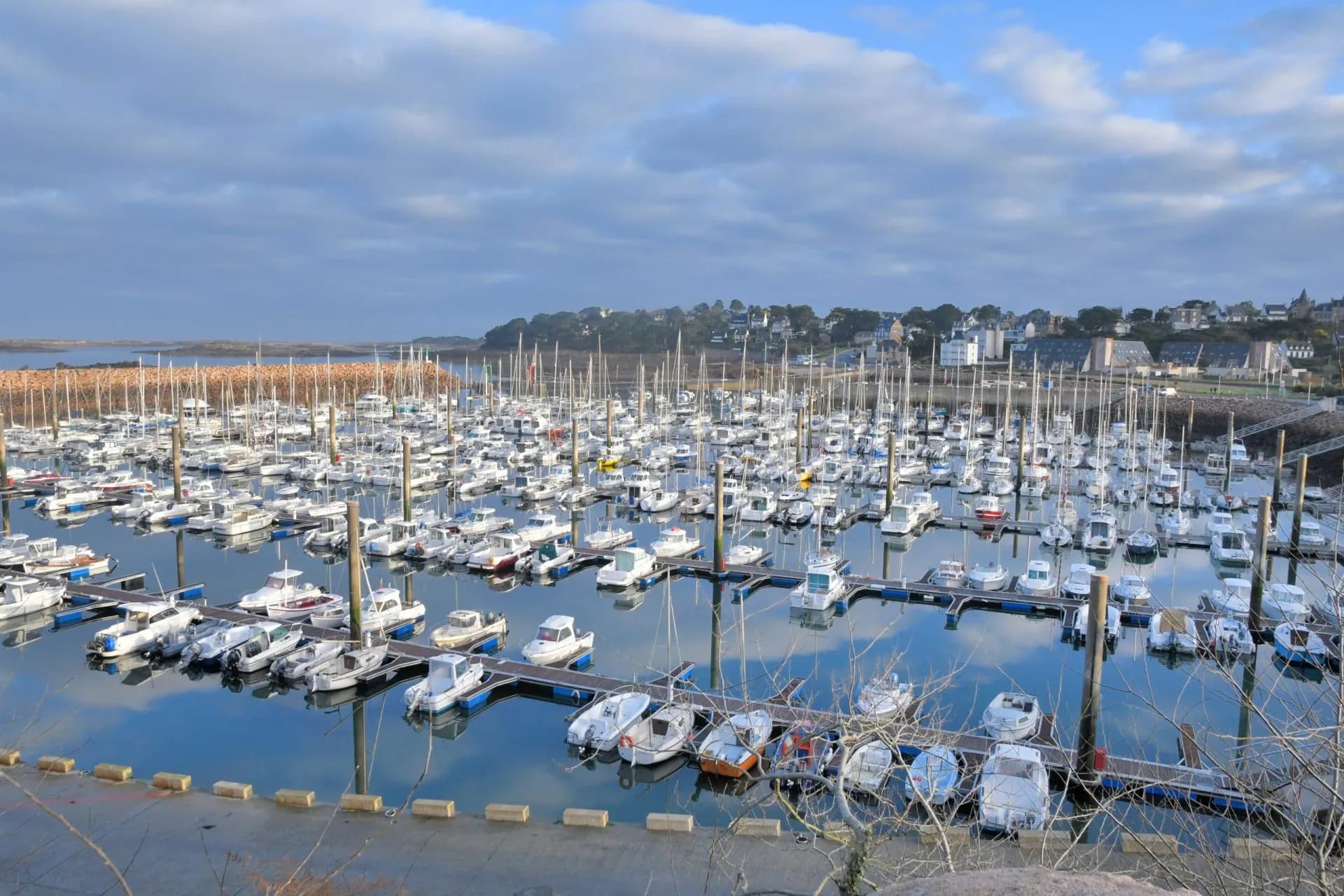Many boats and sailboats moored in the Trebeurden harbour with a town on the distant hillside.