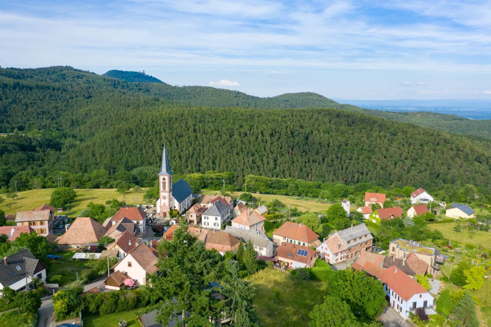 Village with church nestled in green hillsides under a blue sky, Thannenkirch.