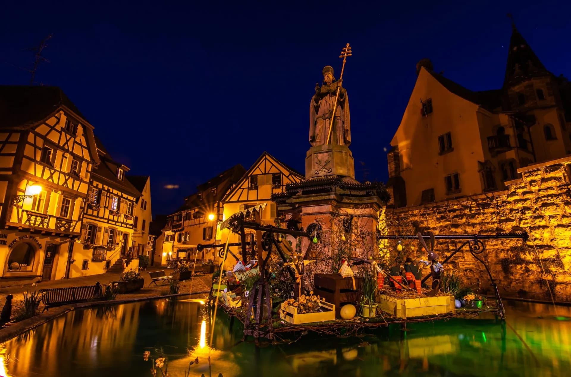 Fountain statue and half-timbered buildings illuminated at night in Eguisheim.