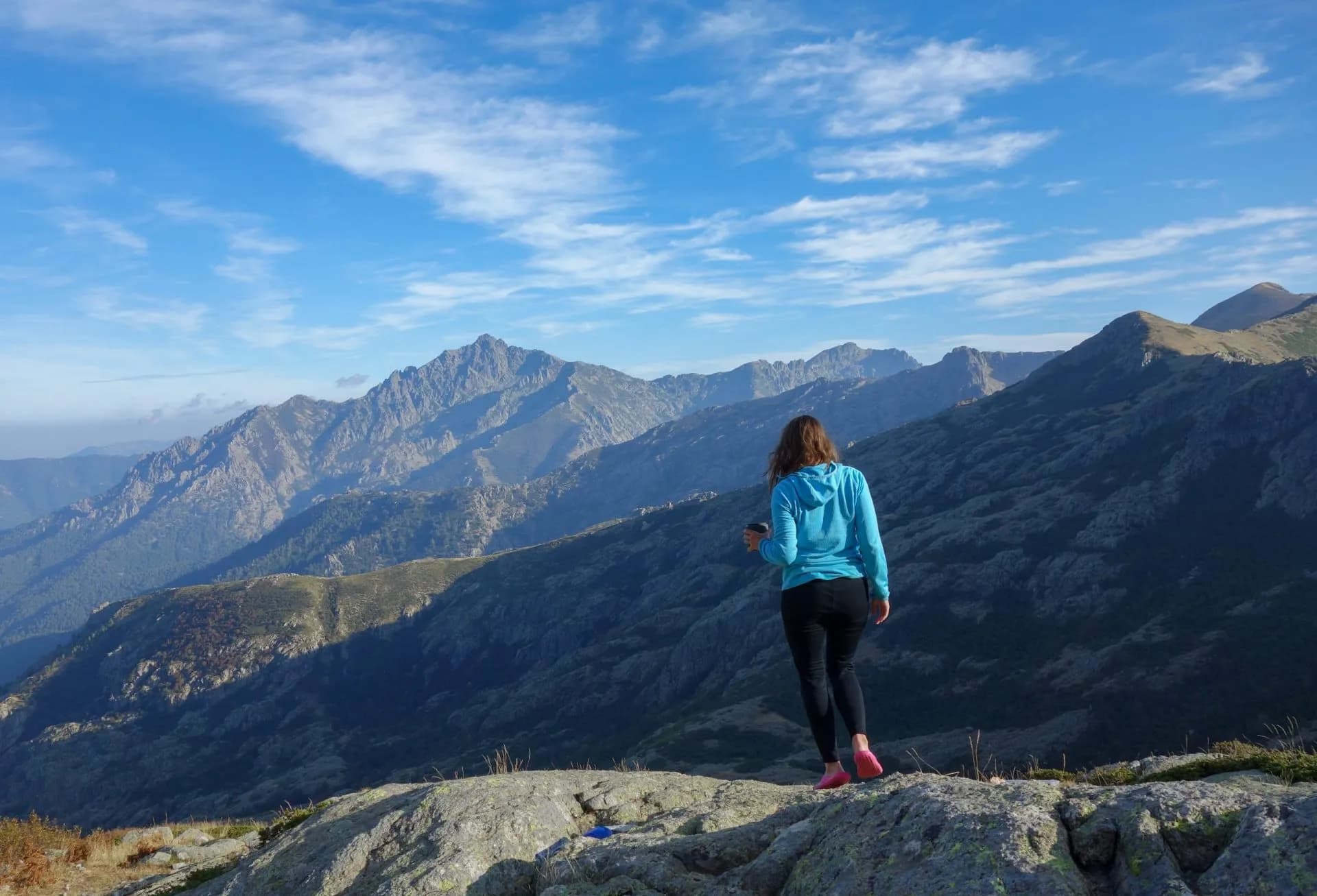 Hiker standing on rocks overlooking rugged mountains under a blue sky in Corsica.