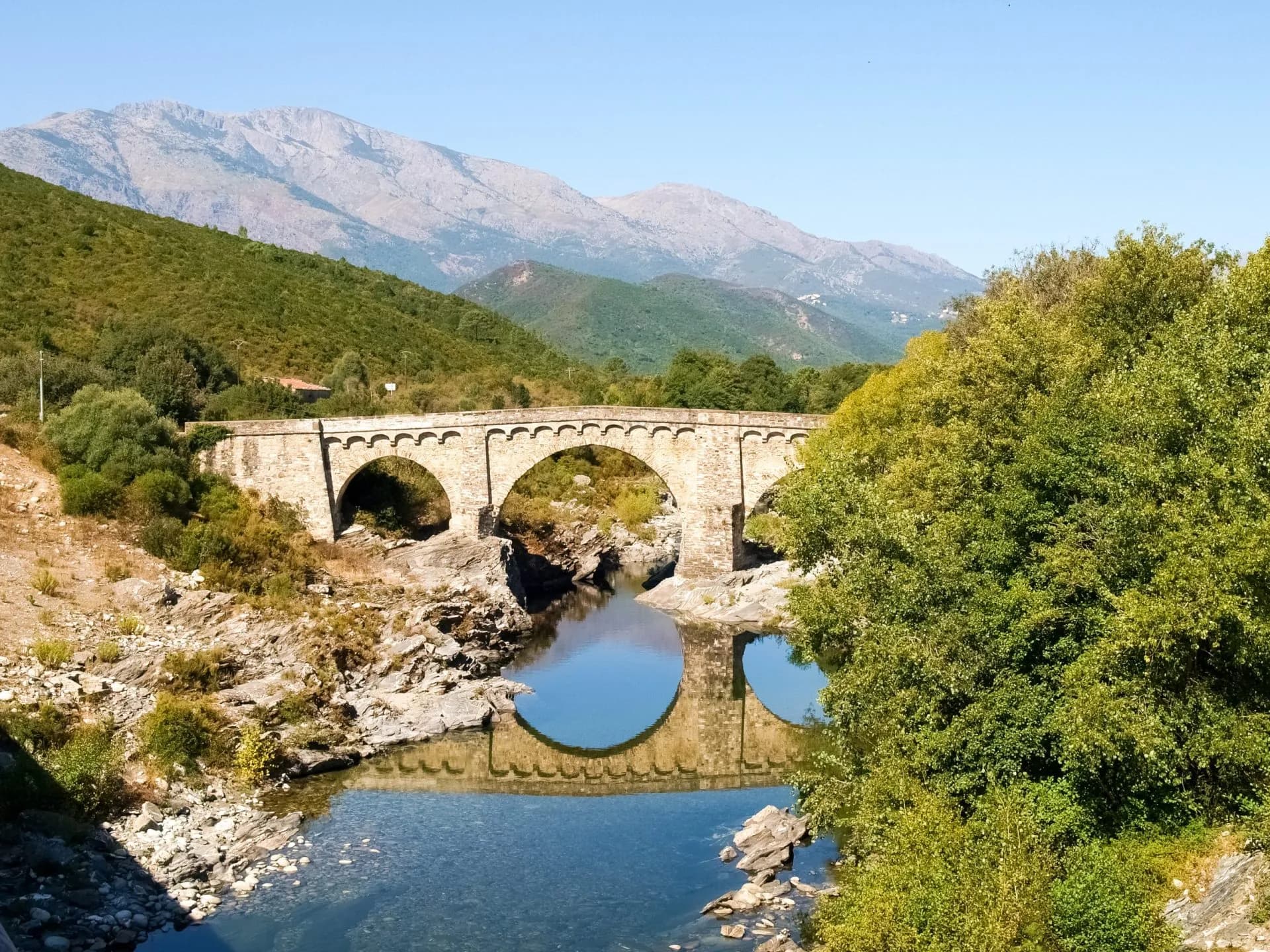 Stone arched bridge over clear river reflecting mountains and green hillsides in Tavignano Gorges.