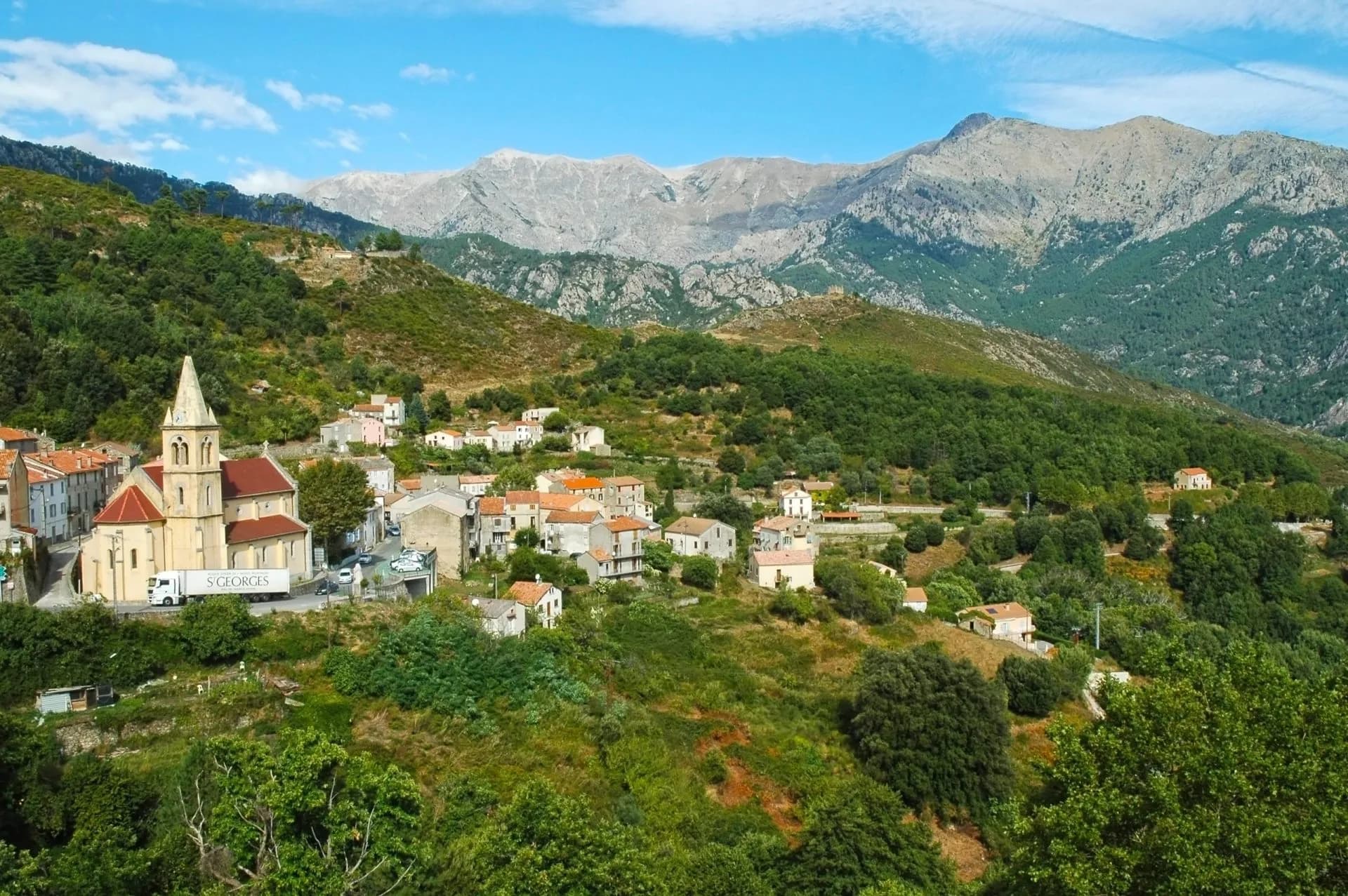 Hillside village with church nestled below rugged, rocky mountains under a blue sky.