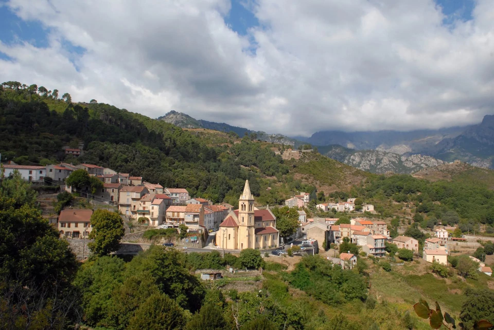 Village with yellow church nestled in lush green mountains under a cloudy sky, Vizzavona.