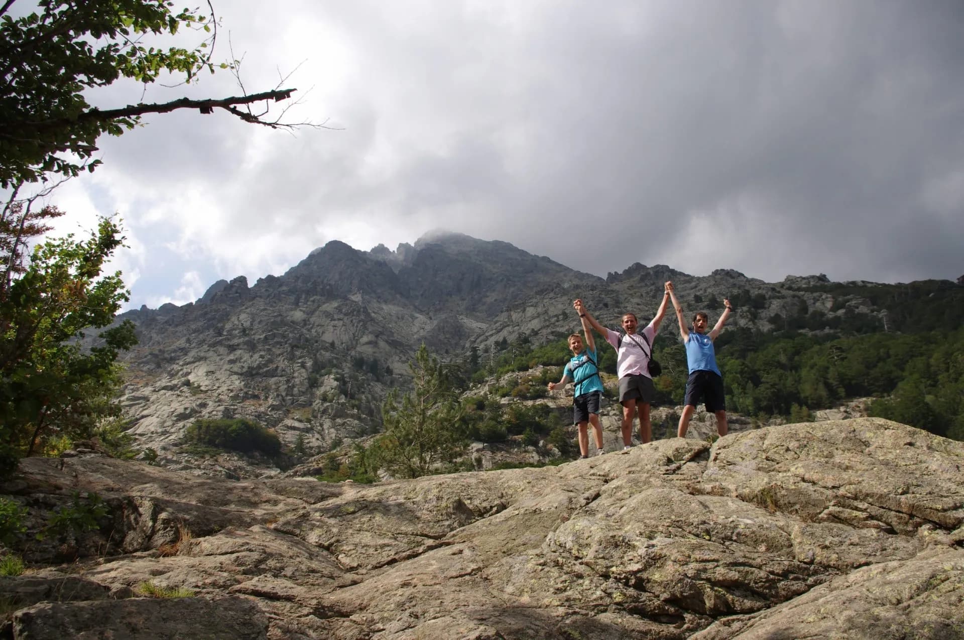 Hikers celebrating on rocky terrain below steep, rugged mountains near Vizzavona.