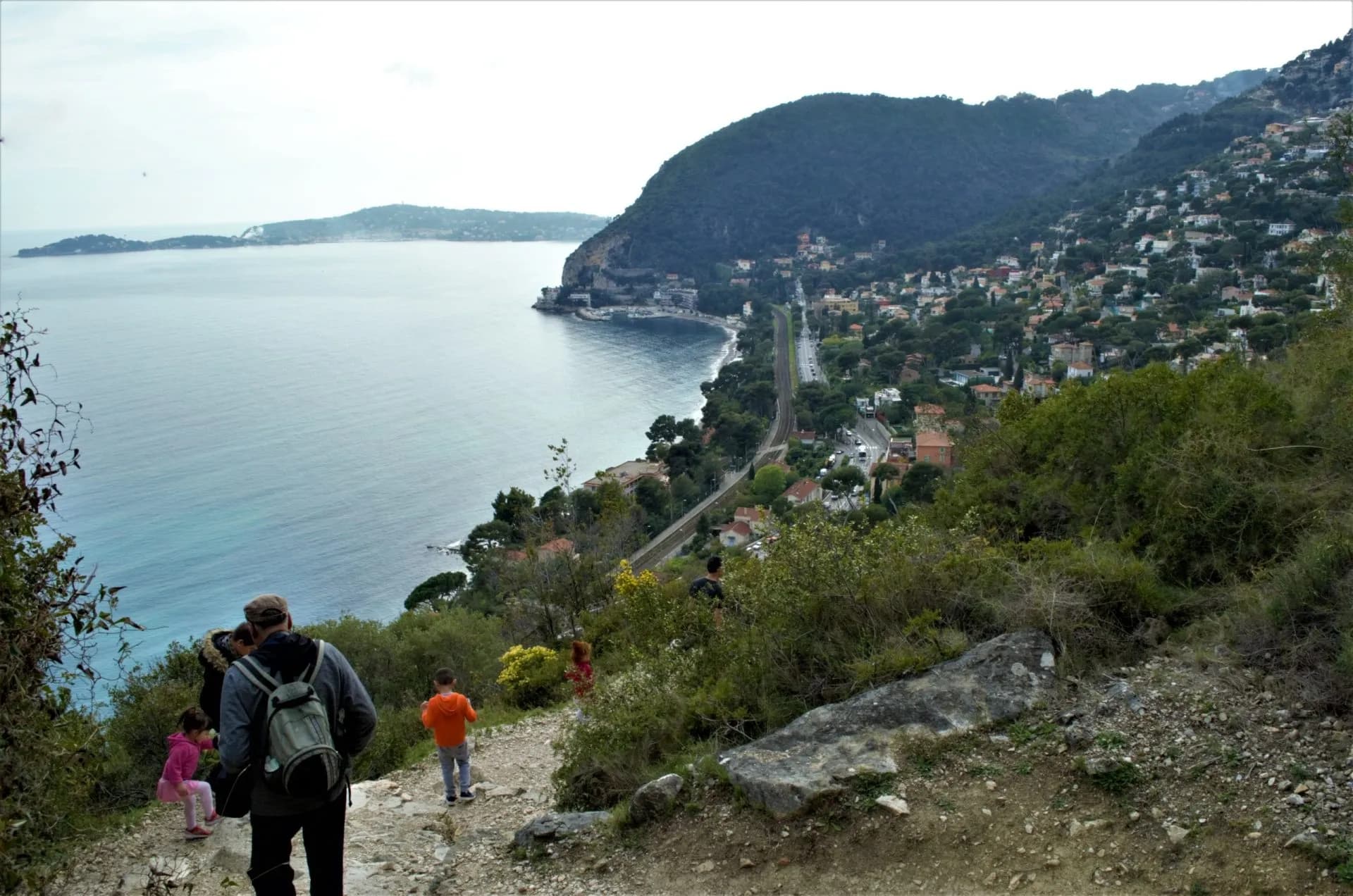 Hikers on dirt path overlooking Mediterranean coastline and hillside town in French Riviera.
