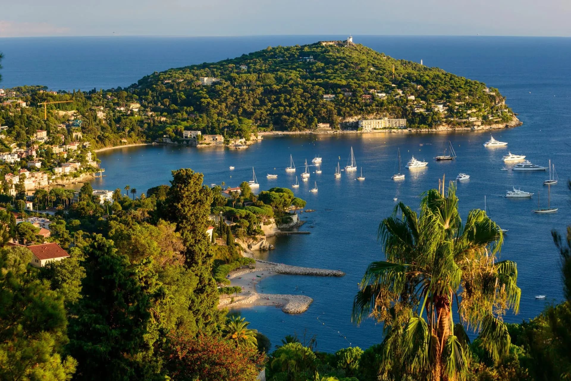Boats anchored in blue bay near lush, green Cap Ferrat peninsula coastline.