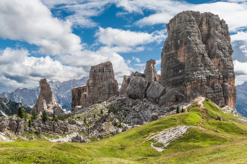 Towering rock formations at Cinque Torri with hikers on a grassy alpine trail under a blue sky.