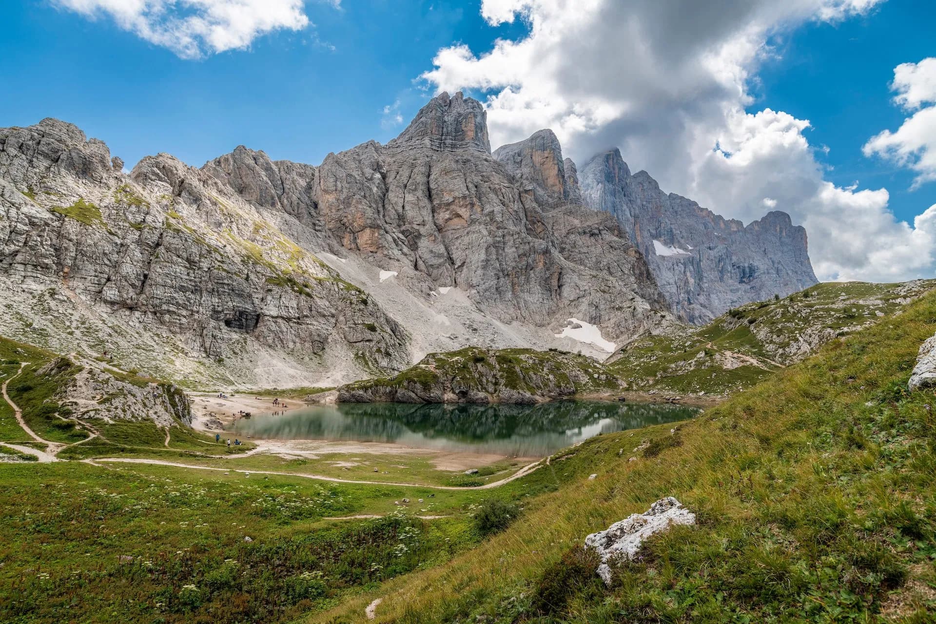 Alpine lake reflecting massive gray mountains under a partly cloudy blue sky, with green meadows.