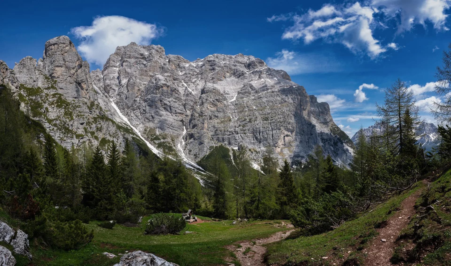 Hiking trail leading to massive gray limestone mountains under a blue, cloudy sky at Passo Duran.