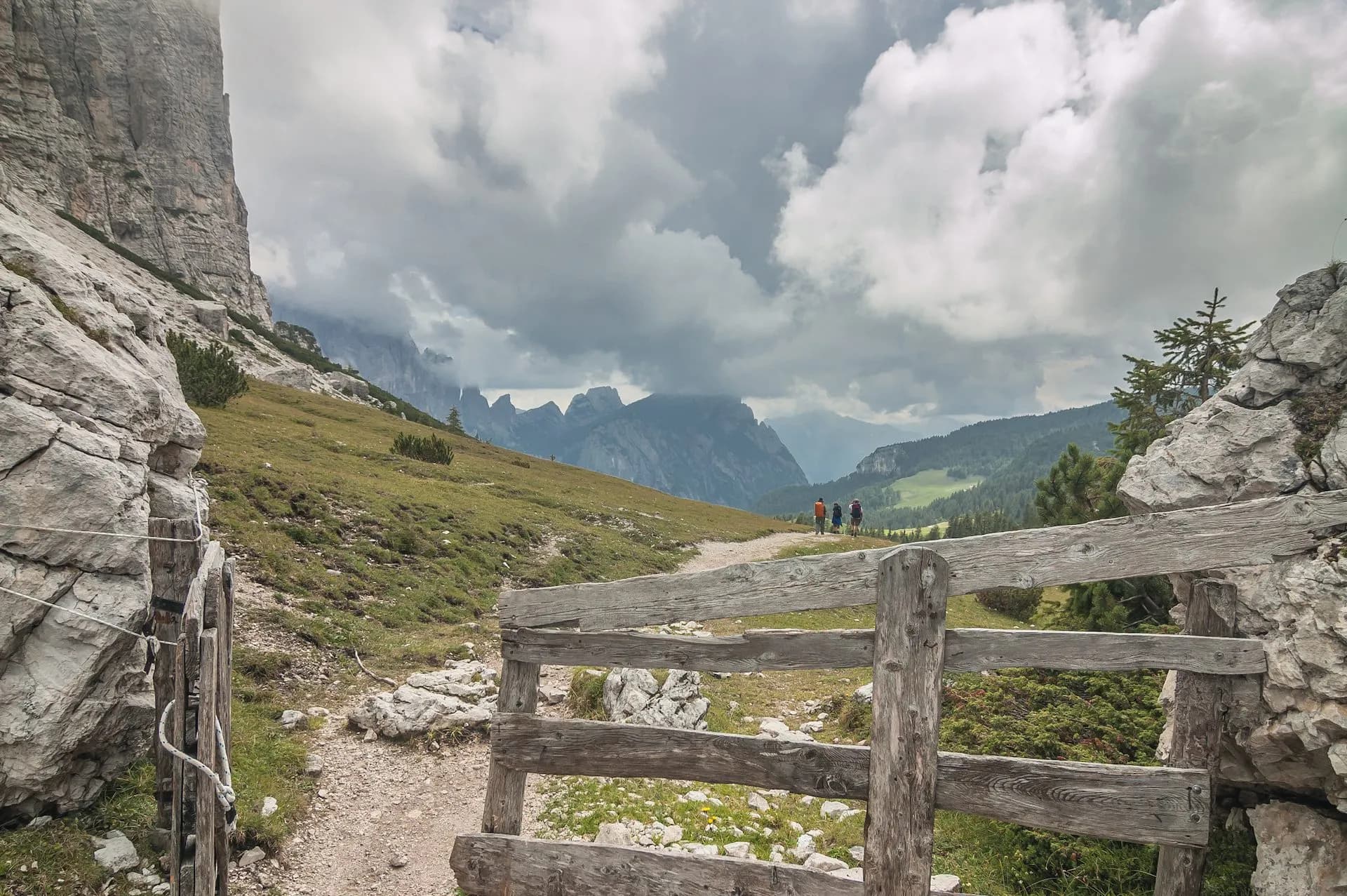 Hikers on dirt path past wooden fence with dramatic mountain range under cloudy sky