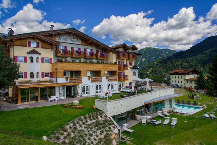 Hotel Renato with balconies, lawn, and pool set against forested mountains under a blue sky.