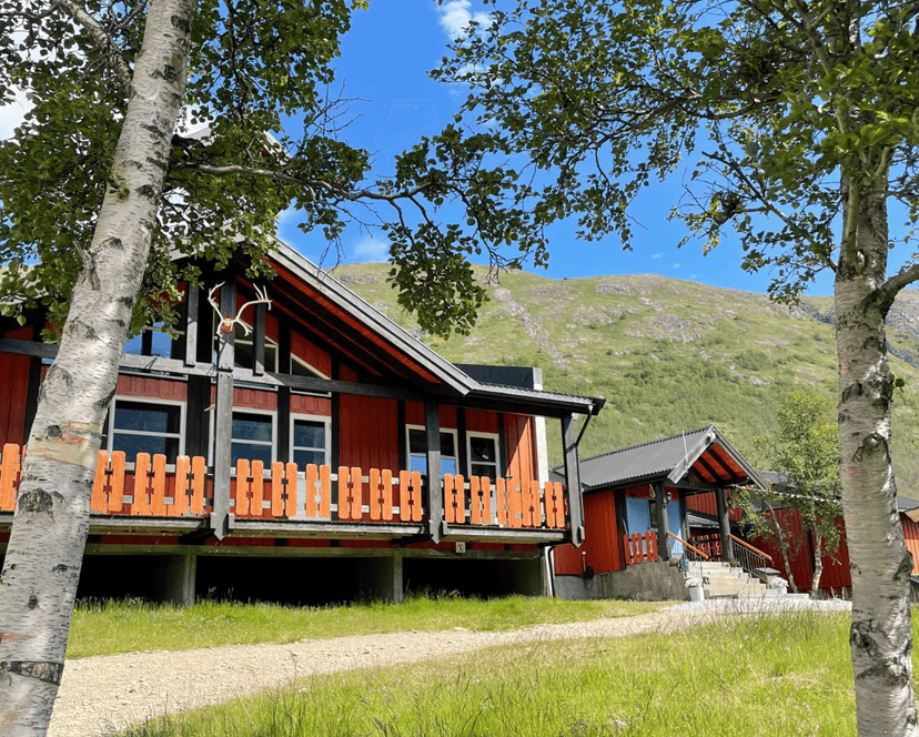 Red wooden tourist cabins with orange railings set against a grassy mountain backdrop under blue sky.