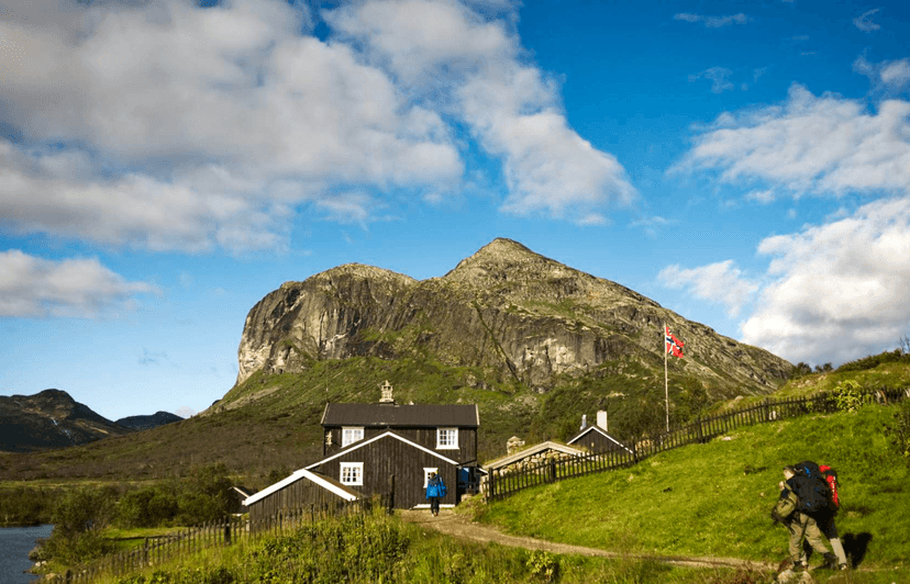 Gjendebu DNT tourist cabin with hikers and Norwegian flag below large green mountain.