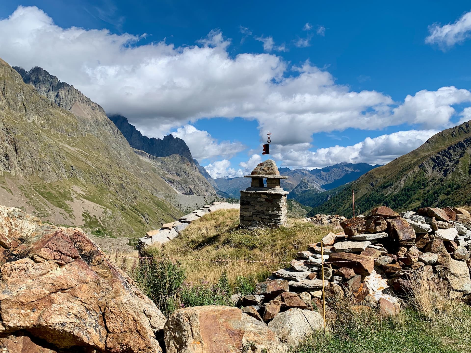 Stone marker with flag on grassy alpine slope near Col de la Seigne, with mountains under blue sky.