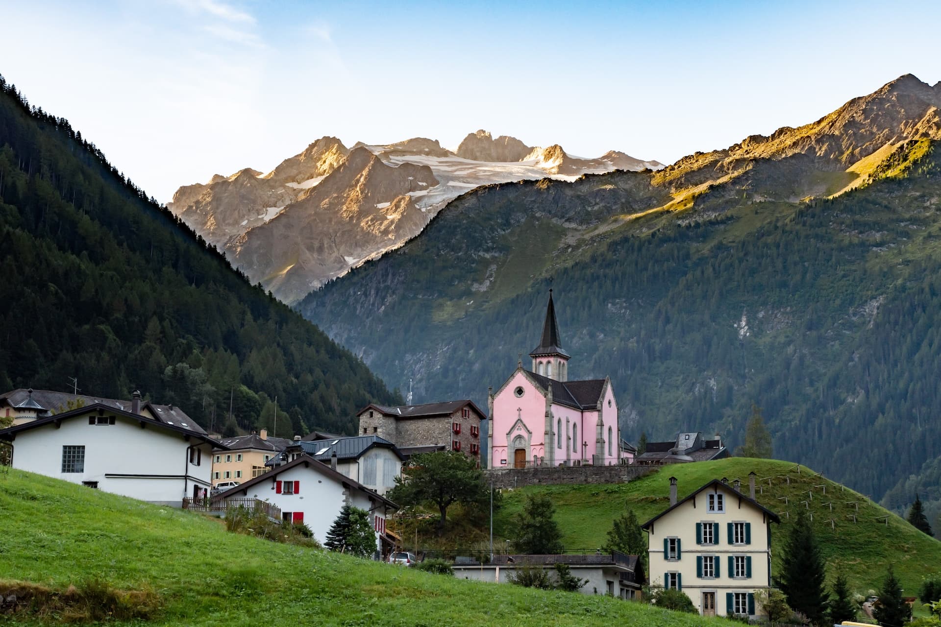 Alpine village with pink church nestled in valley below sunlit, glaciated mountains.