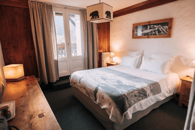 Chalet hotel bedroom with wooden accents, white bedding, and mountain view through balcony doors.