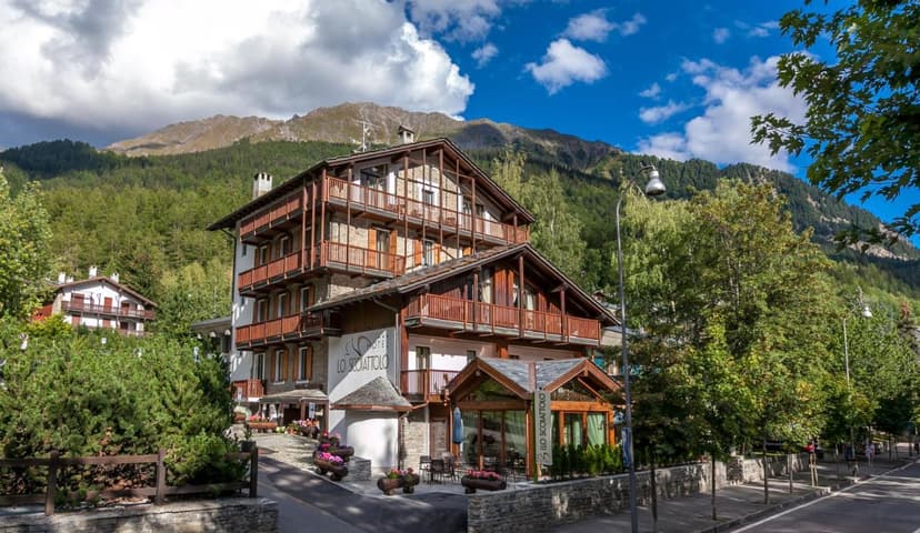 Hotel Lo Scoiattolo with wooden balconies against a backdrop of forested mountains under a blue sky.
