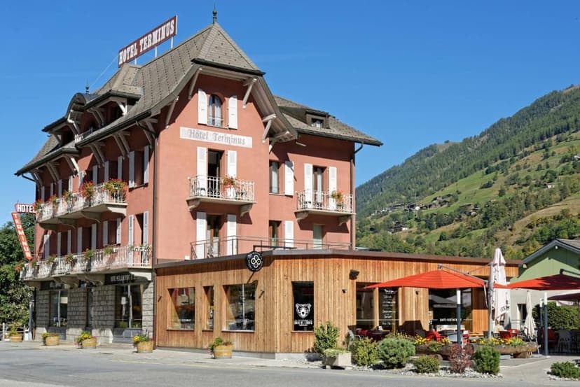 Hotel Terminus building with balconies next to a wooden cafe extension, set against a steep, green mountain slope.