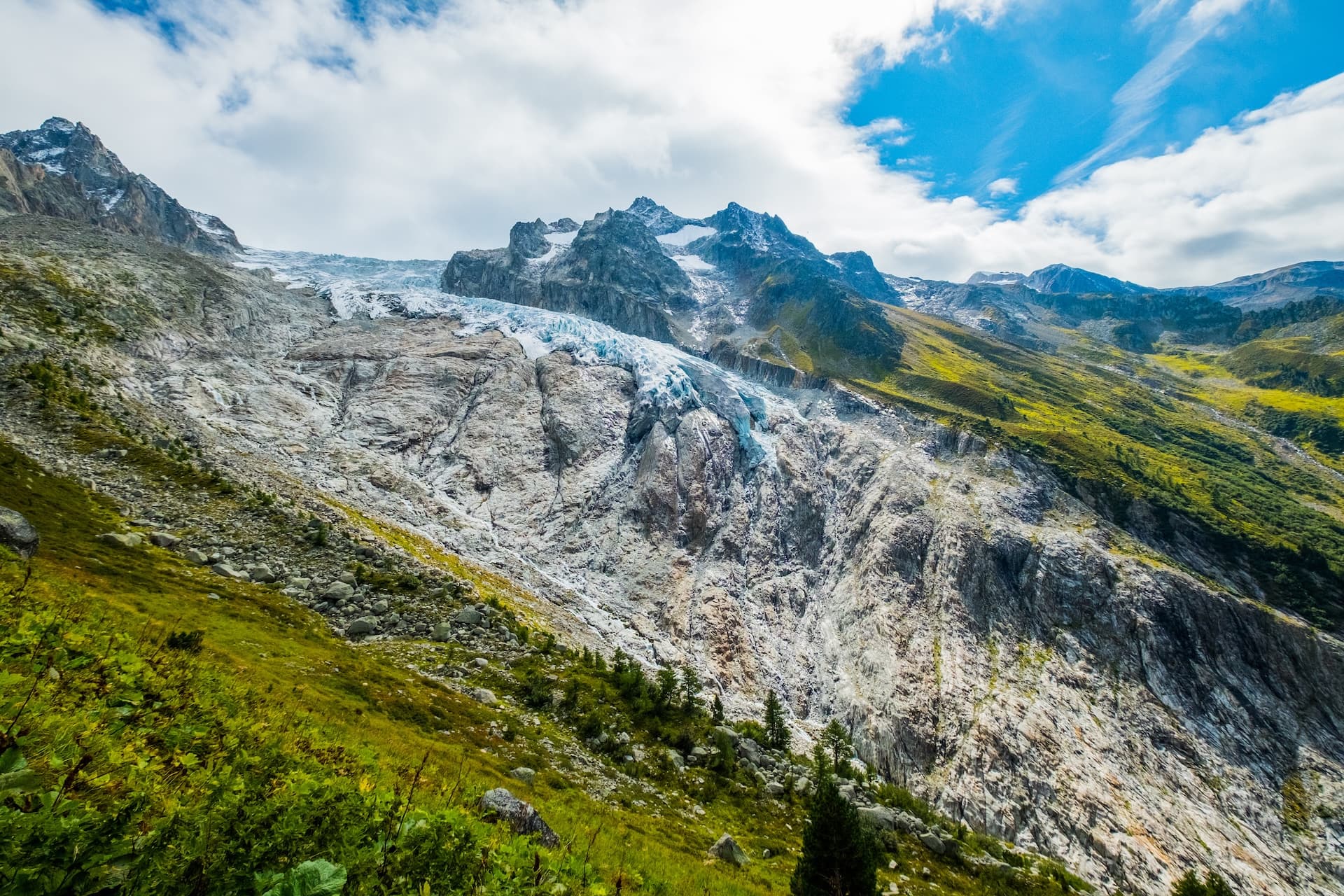 Glacier flowing down rocky mountainside with green slopes under a partly cloudy blue sky.