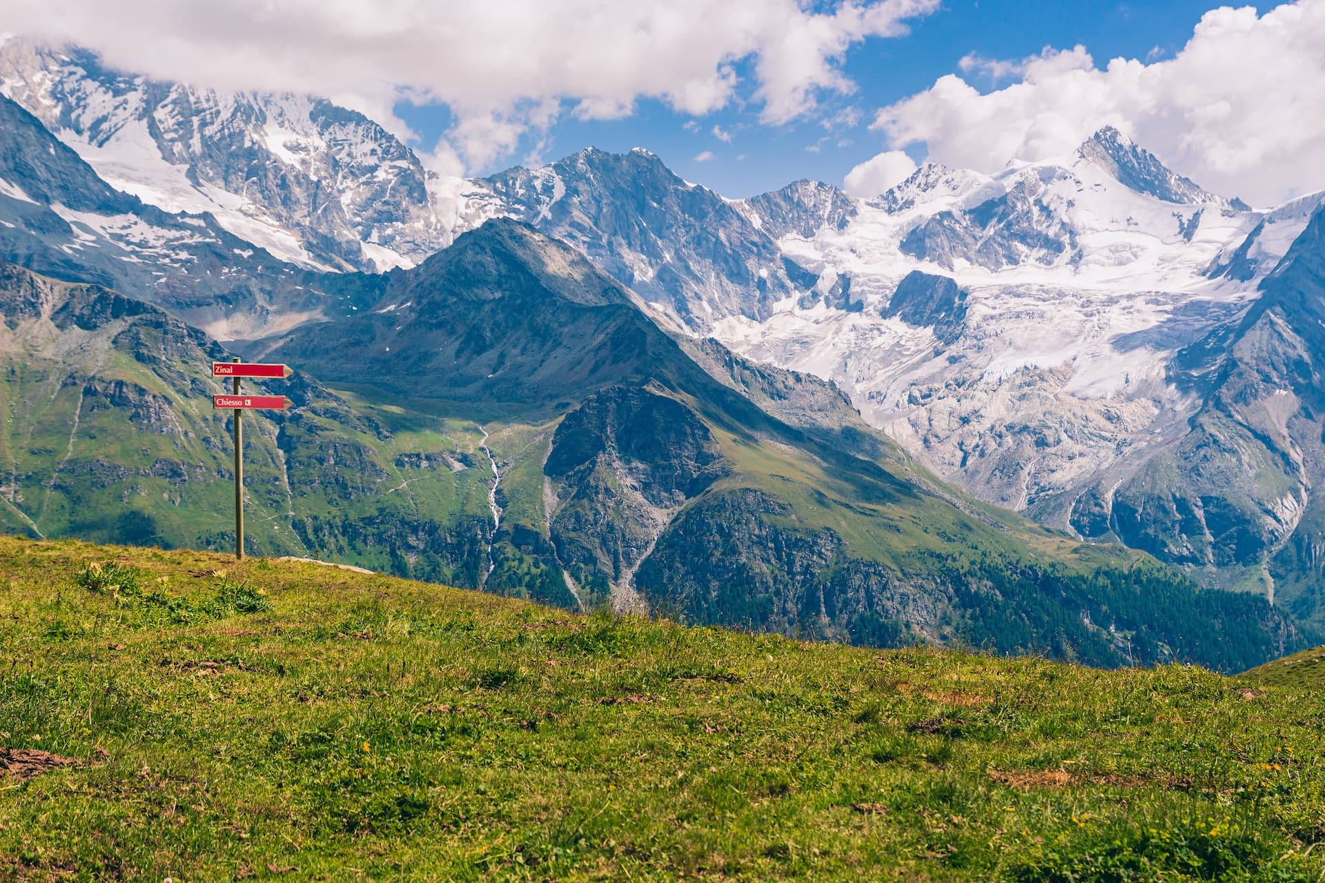 Hiking signpost overlooking green alpine meadow with snow-capped mountains above Saint Luc.