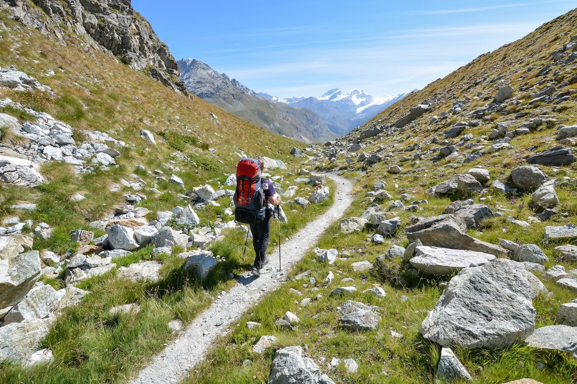 Hiker with backpack trekking on a rocky alpine trail toward snow-capped mountains near Gruben.